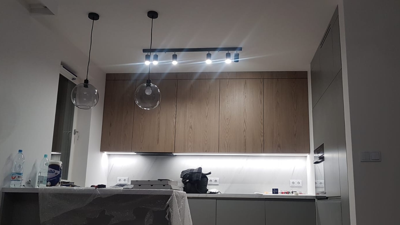 Nowoczesna kuchnia z drewnianymi szafkami, oświetleniem LED podszafkowym i wiszącymi lampami kulami, częściowo zakryta folią ochronną na blacie.