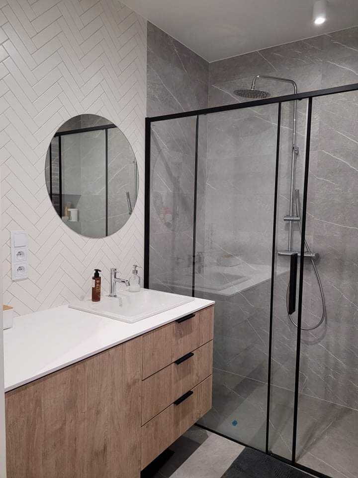 Nowoczesna łazienka z kabiną prysznicową typu walk-in, umywalką nablatową na szafce z drewnopodobnym frontem i okrągłym lustrem na ścianie wyłożonej białymi płytkami ułożonymi w jodełkę.