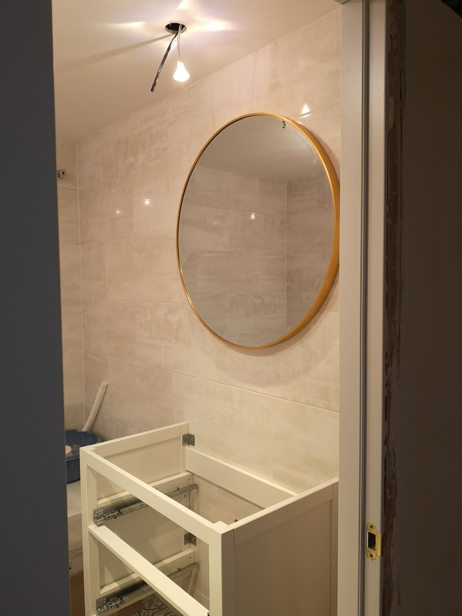 Niedokończona łazienka z beżowymi płytkami, okrągłym lustrem w złotej ramie i niezmontowaną szafką pod umywalkę; widoczne niedokończone wykończenie ościeżnicy i prowizoryczne oświetlenie.