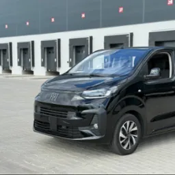 Czarny, nowoczesny bus marki Fiat, zaparkowany na tle magazynu z numerowanymi bramami, z otwartymi drzwiami bocznymi. Widoczne felgi i detale karoserii.