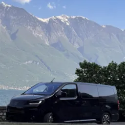 Czarny bus Fiat na tle gór i jeziora, z widocznym fragmentem drogi i niskim murkiem. Pojazd zaparkowany, zarys drzewa po prawej stronie kadru.