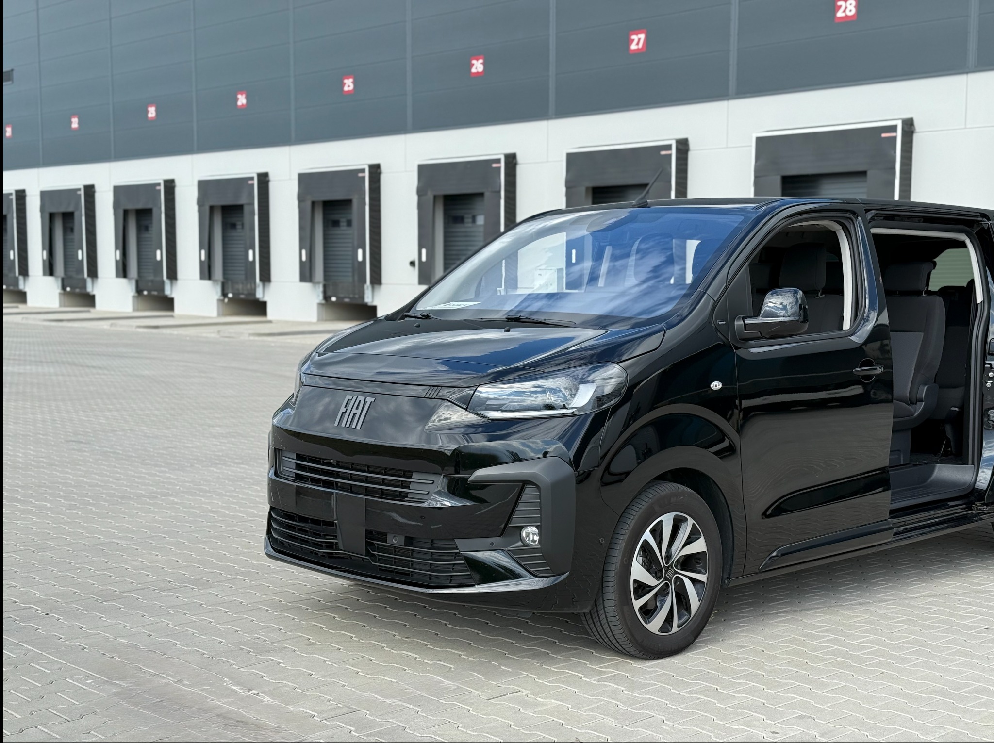 Czarny, nowoczesny bus marki Fiat, zaparkowany na tle magazynu z numerowanymi bramami, z otwartymi drzwiami bocznymi. Widoczne felgi i detale karoserii.
