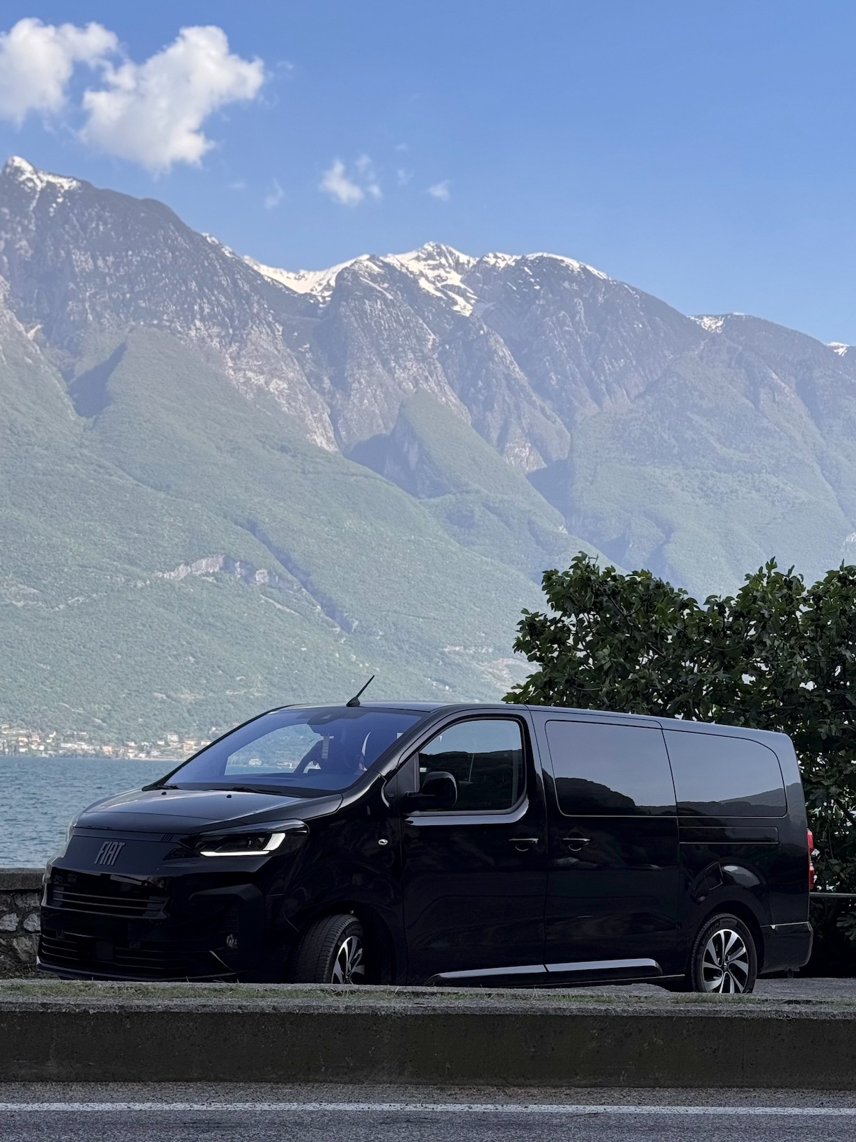 Czarny bus Fiat na tle gór i jeziora, z widocznym fragmentem drogi i niskim murkiem. Pojazd zaparkowany, zarys drzewa po prawej stronie kadru.