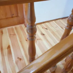 Drewniane schody z balustradą z toczonymi tralkami. Jasna podłoga z wyraźnym usłojeniem. Widoczny fragment drzwi w tle.