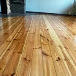 Błyszcząca, odnowiona podłoga z desek w odcieniach miodowego brązu, z widocznymi słojami drewna. W tle ściana w kolorze gołębim i fragment kominka.