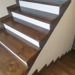 Odnowione drewniane schody z brązowymi stopniami i białymi podstopnicami, widoczne z dołu, z wyraźnym rysunkiem drewna i połyskiem lakieru.