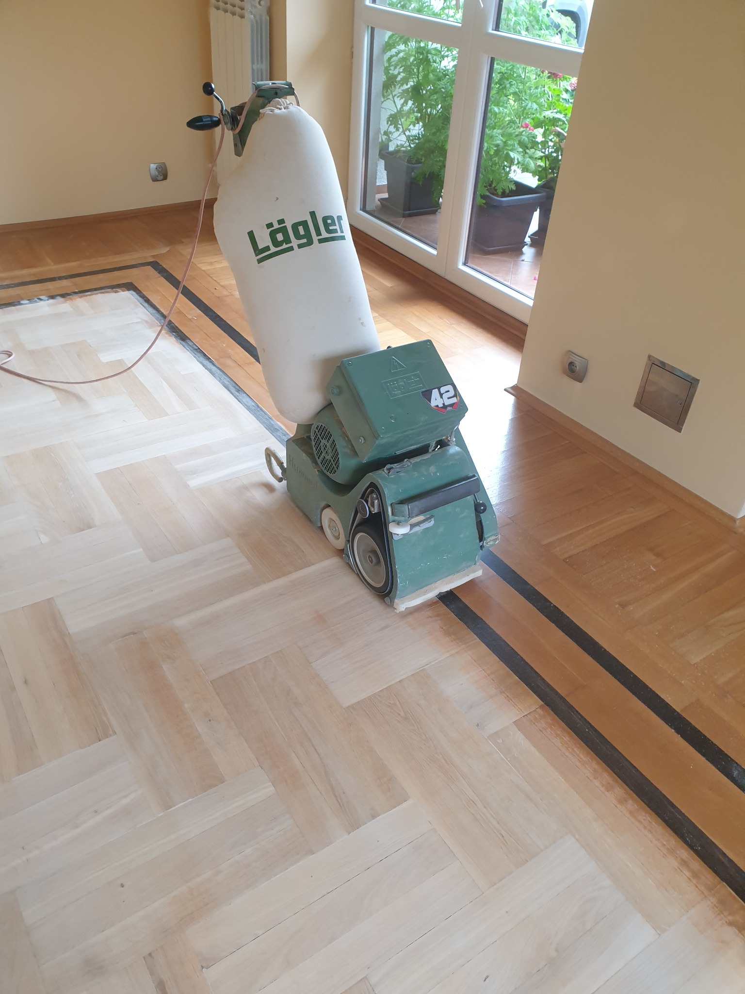 Parkiet w jodełkę po cyklinowaniu, widoczna maszyna marki Läglera. Część podłogi wycyklinowana, reszta w ciemnym kolorze. Okno balkonowe w tle.