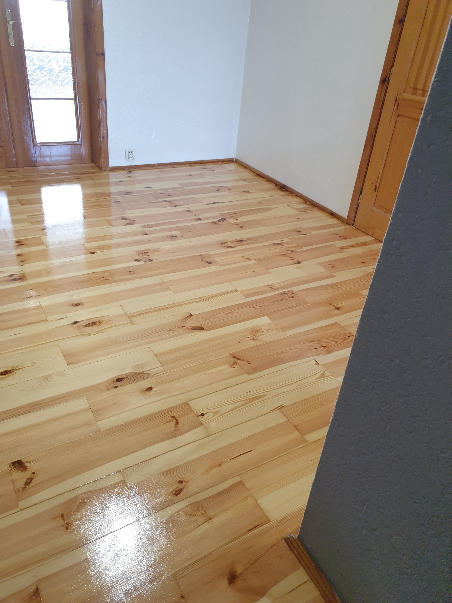 Jasne, lakierowane deski podłogowe z widocznym usłojeniem, świeżo po renowacji. Fragment ściany i drzwi w tle. Podłoga odbija światło.