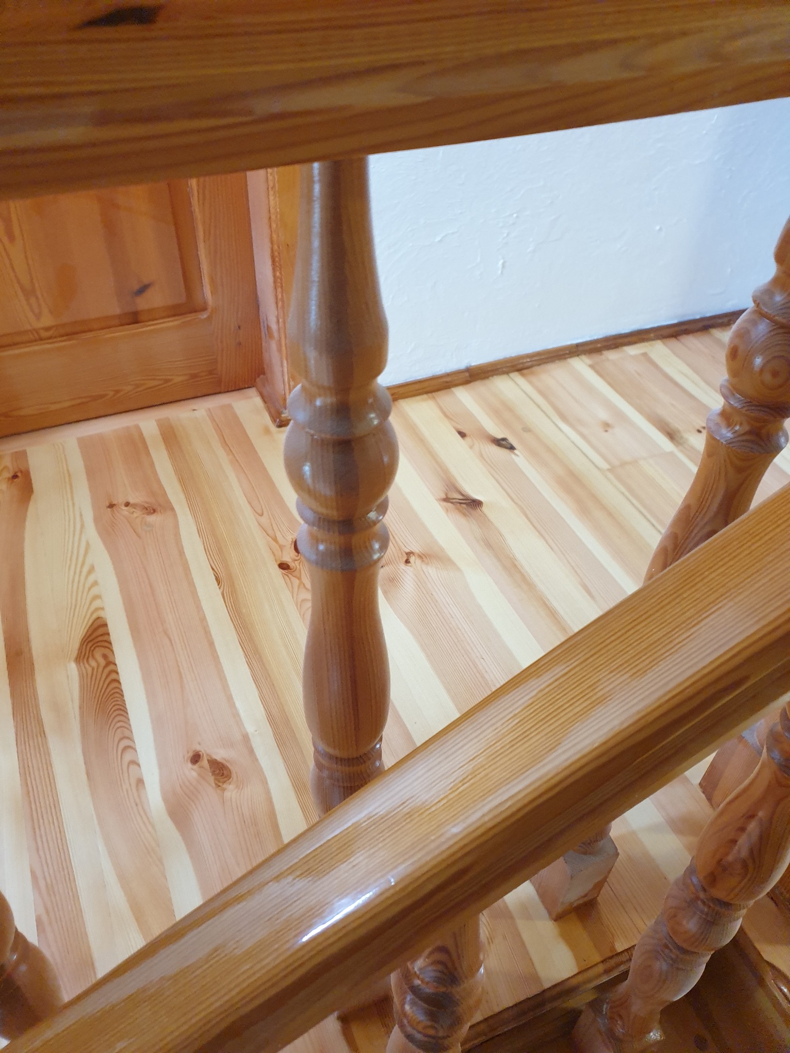 Drewniane schody z balustradą z toczonymi tralkami. Jasna podłoga z wyraźnym usłojeniem. Widoczny fragment drzwi w tle.