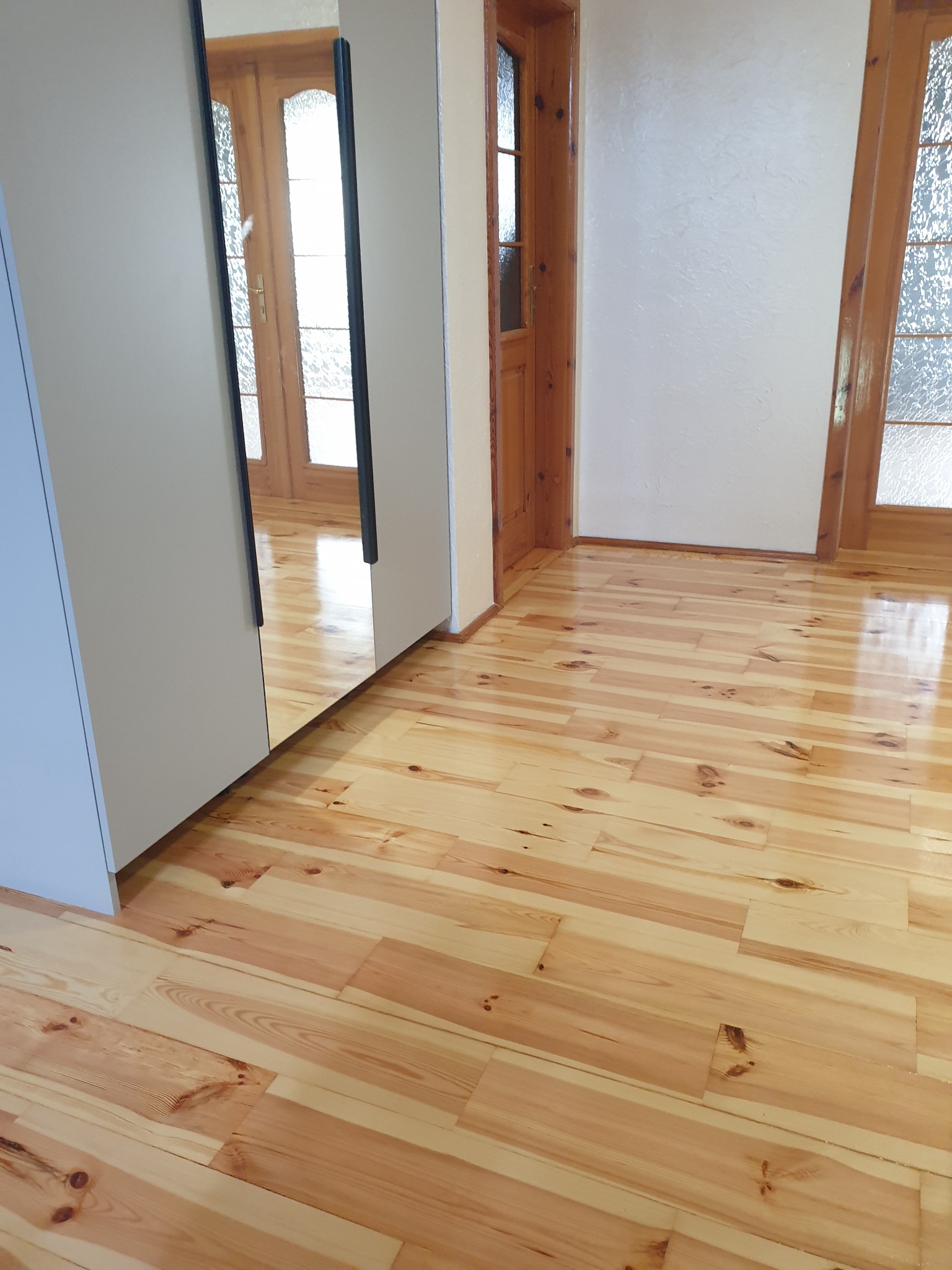 Jasna, odświeżona podłoga z desek, widoczna z perspektywy narożnika pokoju, z fragmentem szafy z lustrem i drewnianymi drzwiami w tle. Parkiet po renowacji.