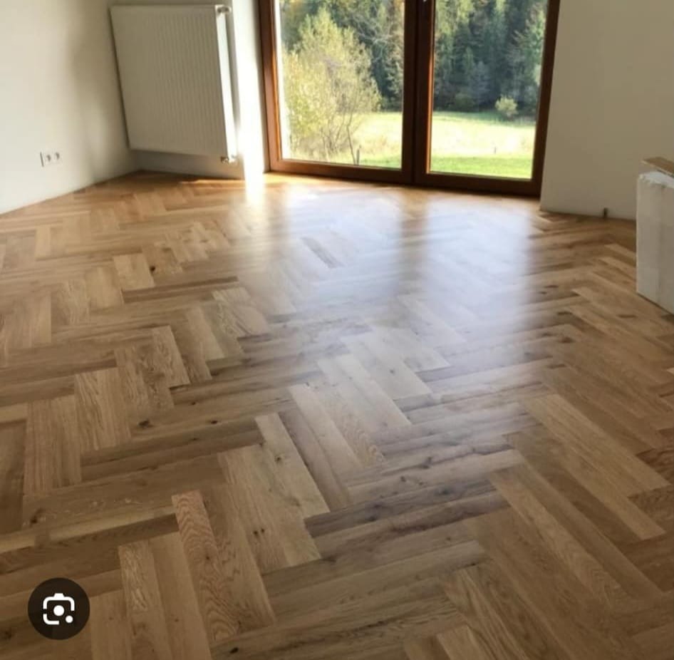 Jasny parkiet ułożony we wzór jodełki francuskiej w przestronnym pokoju z dużym oknem i widokiem na zieleń. Naturalne światło podkreśla strukturę drewna.