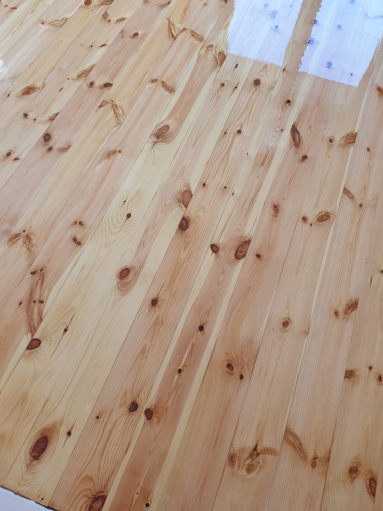 Jasne, drewniane deski podłogowe, częściowo pokryte lakierem, ukazujące naturalny wzór słojów i sęków. Ukośny kadr podkreśla strukturę drewna.