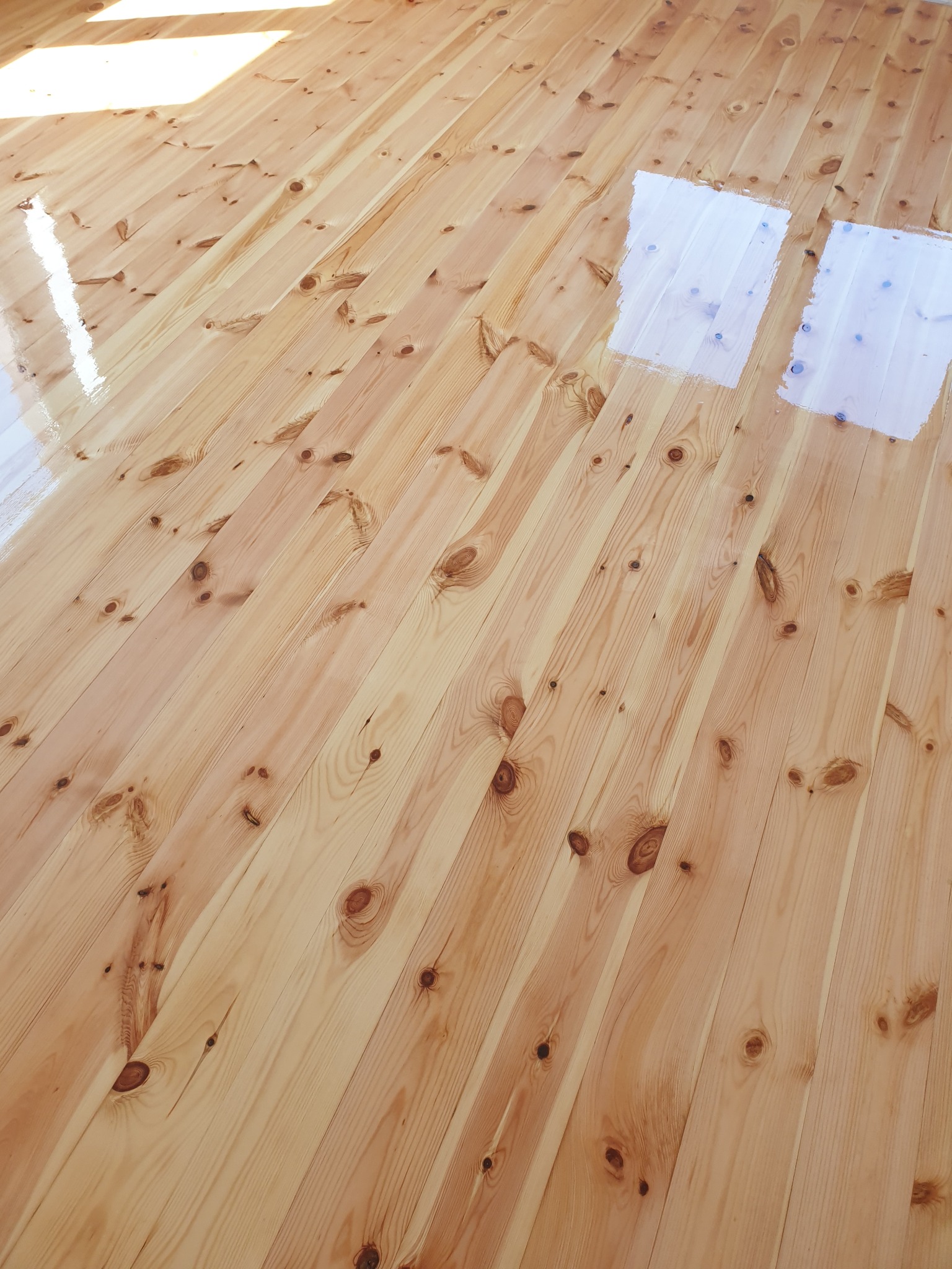 Jasna podłoga z desek po częściowym malowaniu, widoczne fragmenty surowego drewna i świeżo nałożona biała farba, diagonalne ułożenie desek, oświetlenie naturalne.