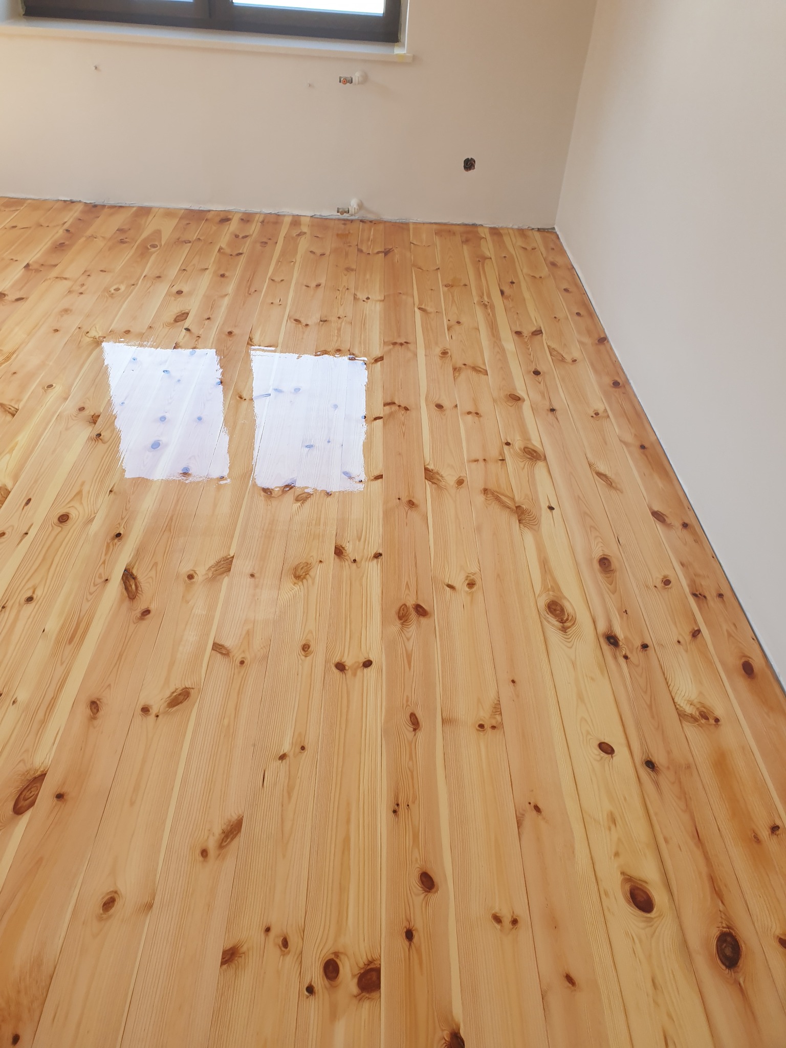 Jasna, drewniana podłoga z widocznymi, świeżo nałożonymi dwiema warstwami lakieru. Widok z góry na fragment wnętrza z oknem w górnej części kadru.