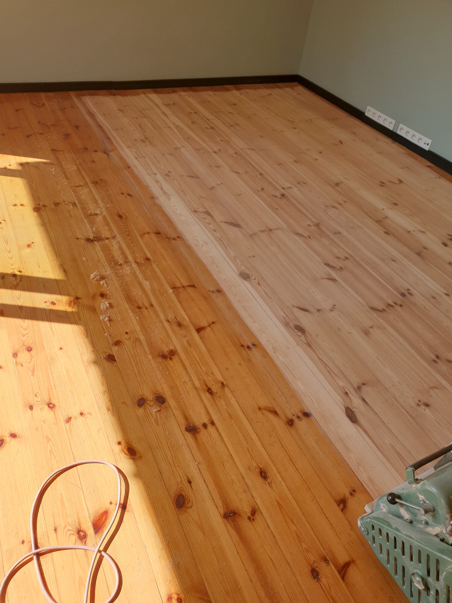 Częściowo odnowiona sosnowa podłoga z widocznym sprzętem do cyklinowania. Kontrast między surowym drewnem a odnowioną powierzchnią. Kabel na pierwszym planie.