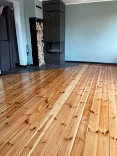 Wypolerowana, lakierowana podłoga z desek w Koziegłowach, z widocznym kominkiem w tle. Parkiet po cyklinowaniu z wyraźnym połyskiem, podkreślającym usłojenie drewna.