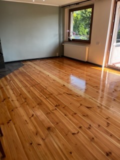 Błyszcząca, odnowiona podłoga z desek w jasnym salonie z oknem. Widoczne refleksy światła na lakierowanej powierzchni. Ściany w odcieniach szarości i beżu.