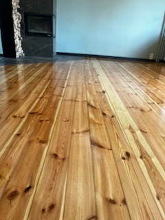 Błyszcząca, odnowiona podłoga z desek w odcieniach miodowego brązu, z widocznymi słojami drewna. W tle ściana w kolorze gołębim i fragment kominka.