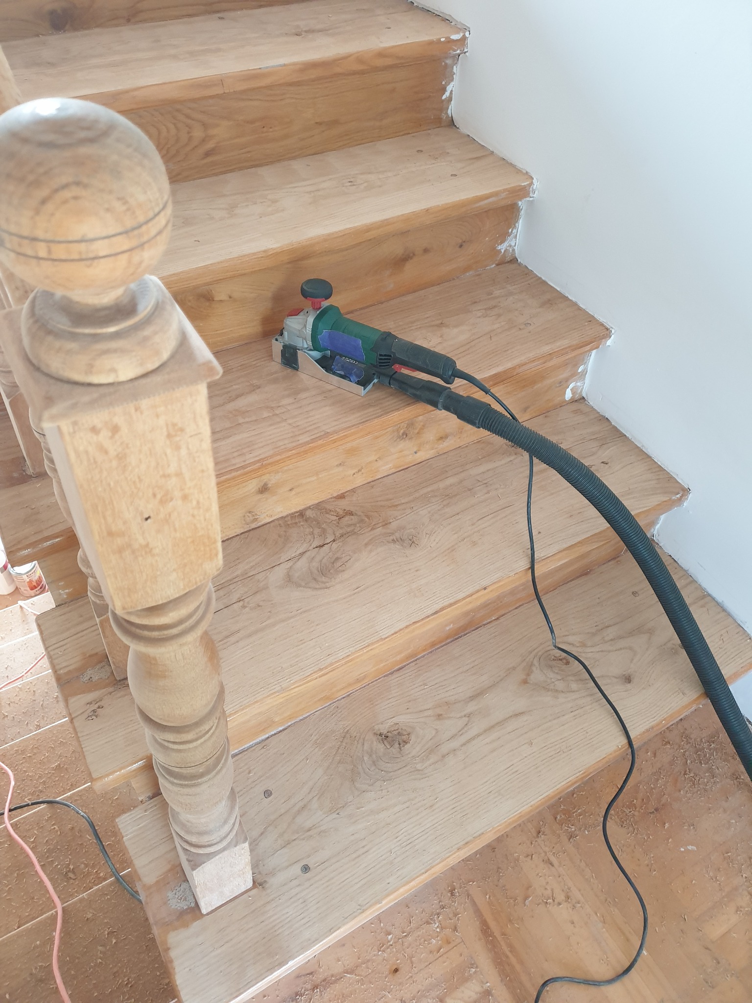 Renowacja drewnianych schodów z użyciem szlifierki kątowej z odkurzaczem. Widoczne drewniane stopnie i balustrada w trakcie obróbki.