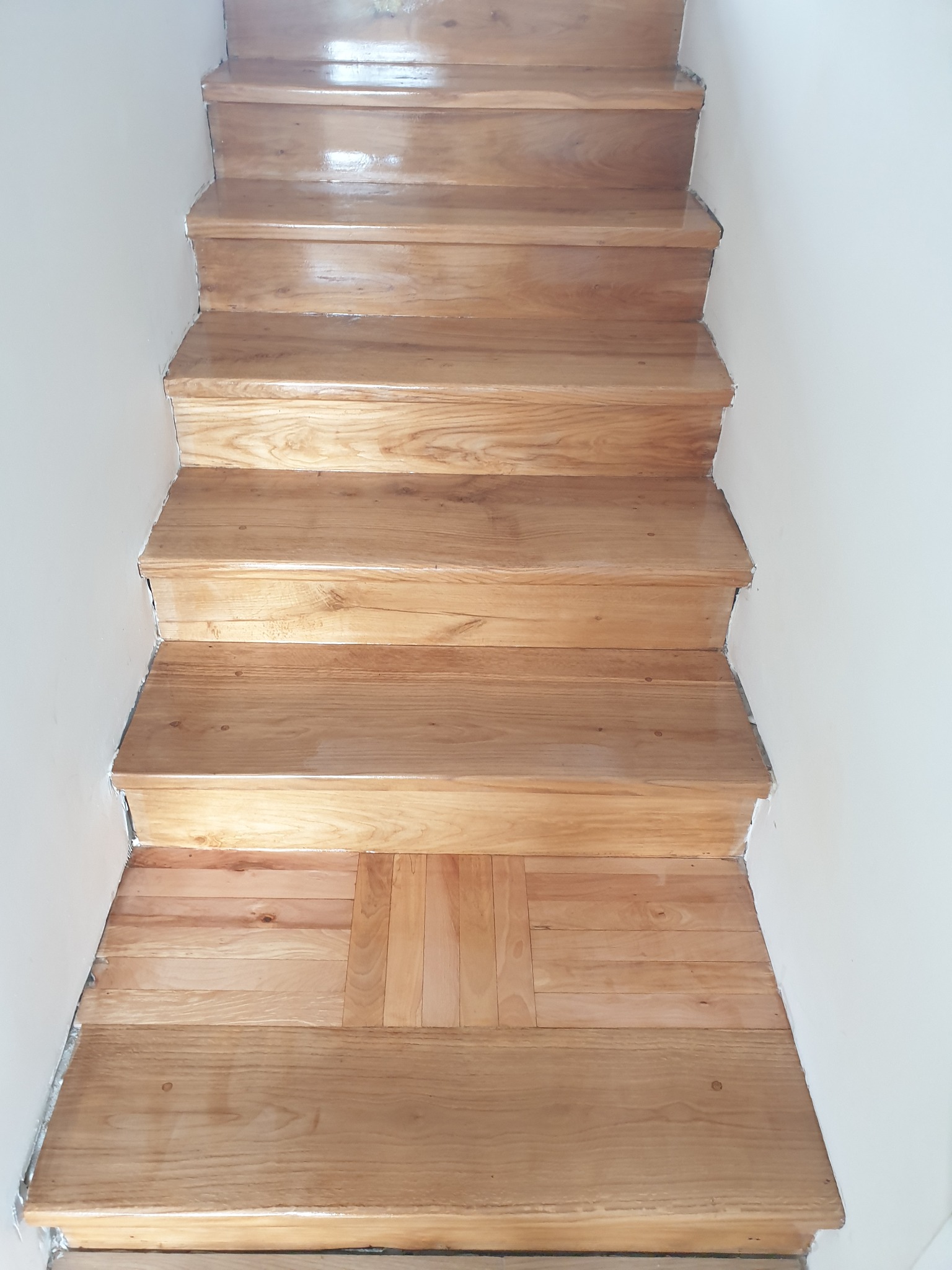 Odnowione, drewniane schody z jasnego drewna, prowadzące w górę, z widocznymi detalami wykończenia przy białej ścianie. Parkiet na podeście ułożony w jodełkę.