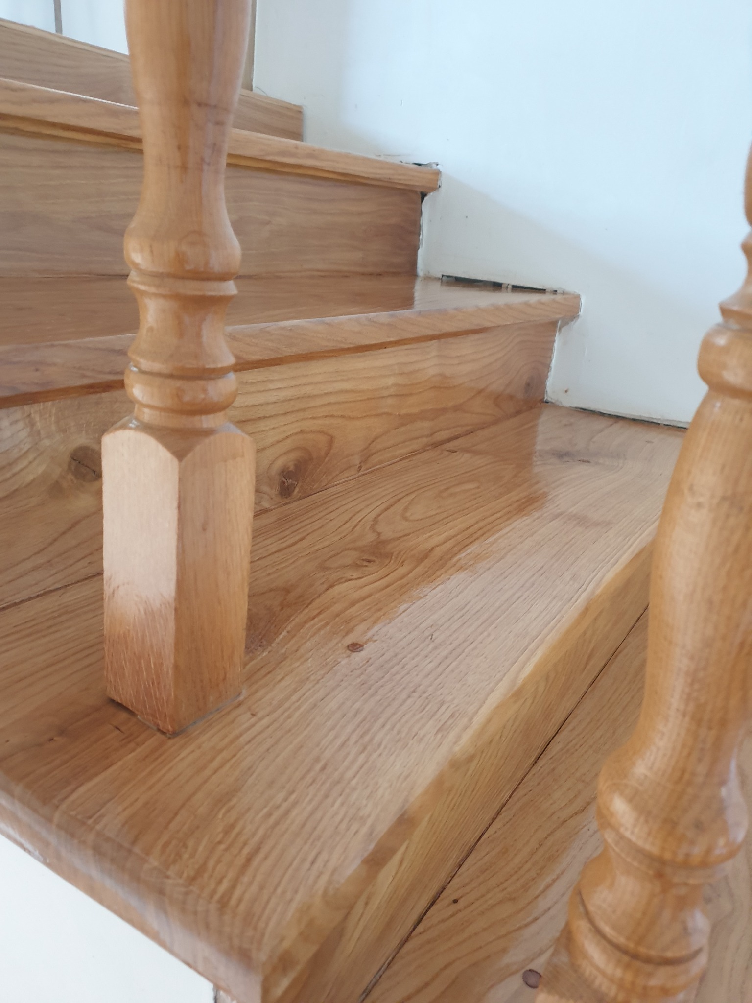 Polakierowane drewniane schody z ozdobnymi tralkami, widok z dołu. Jasne drewno o widocznym usłojeniu, połyskująca powierzchnia, fragment białej ściany w tle.