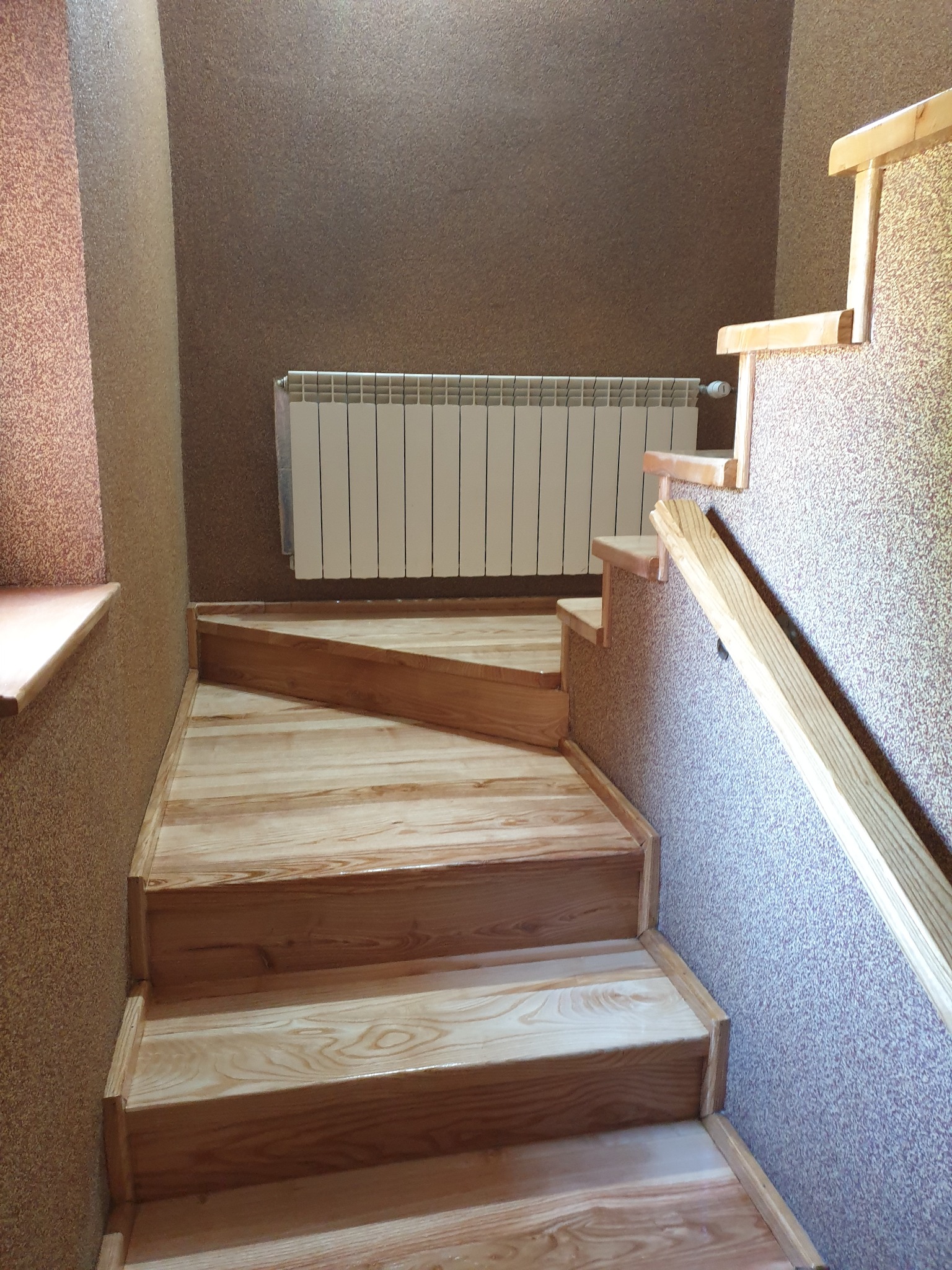 Jasne, drewniane schody z poręczą w domu. Widoczny grzejnik na ścianie o fakturze korka. Ujęcie od dołu schodów, podkreślające detale wykończenia.