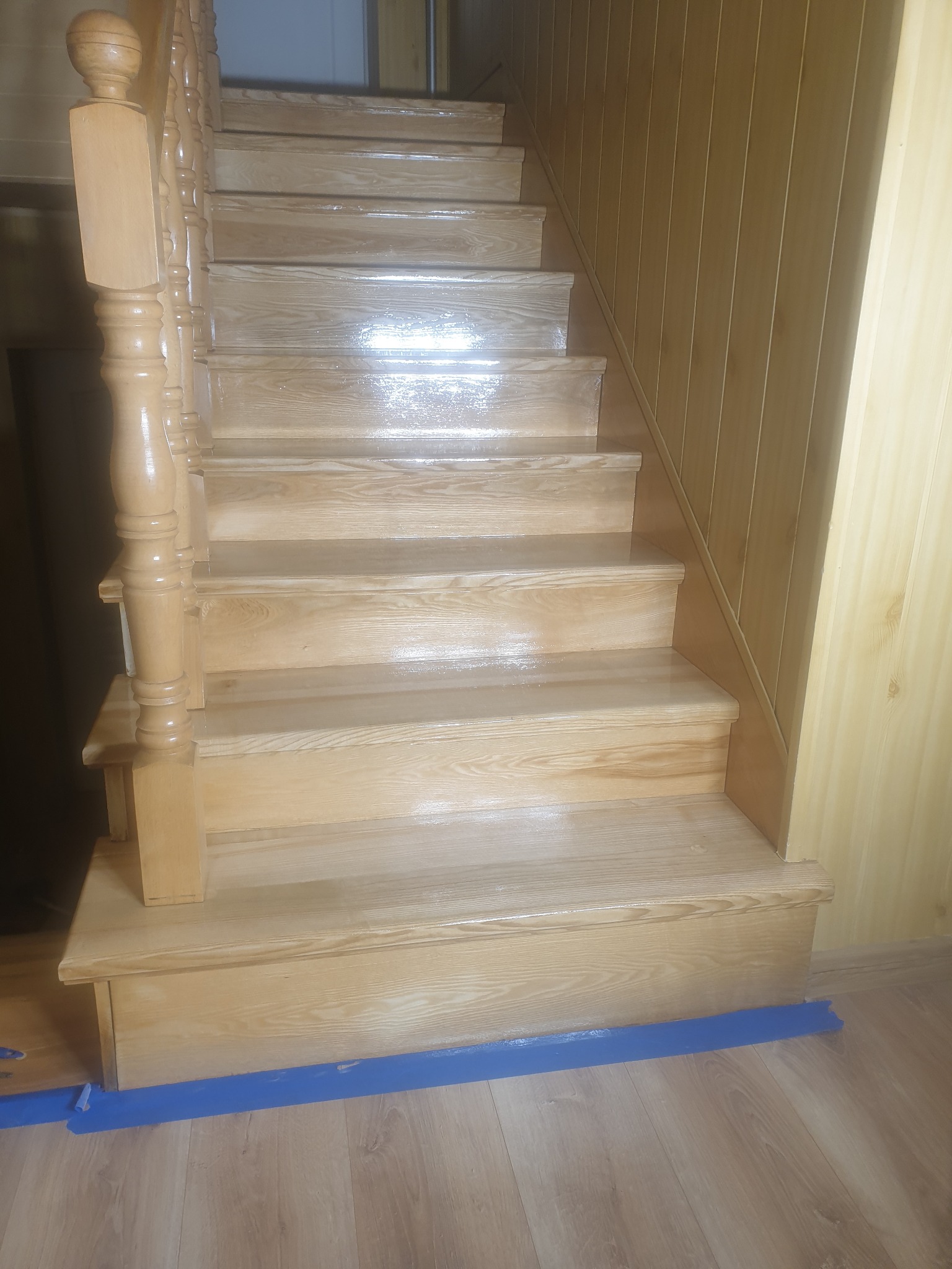Drewniane schody po renowacji, zabezpieczone niebieską taśmą malarską przy podłodze. Widoczny połysk lakieru i odnowiona balustrada.