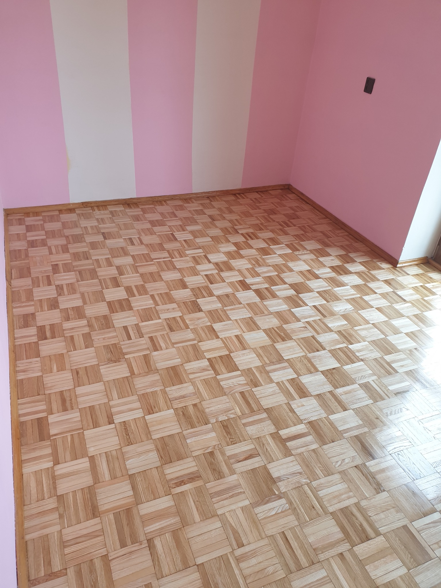 Ukończona podłoga z parkietu w kwadraty w pokoju z różowo-białymi pasami na ścianie i brązową listwą przypodłogową. Widok na podłogę w perspektywie.