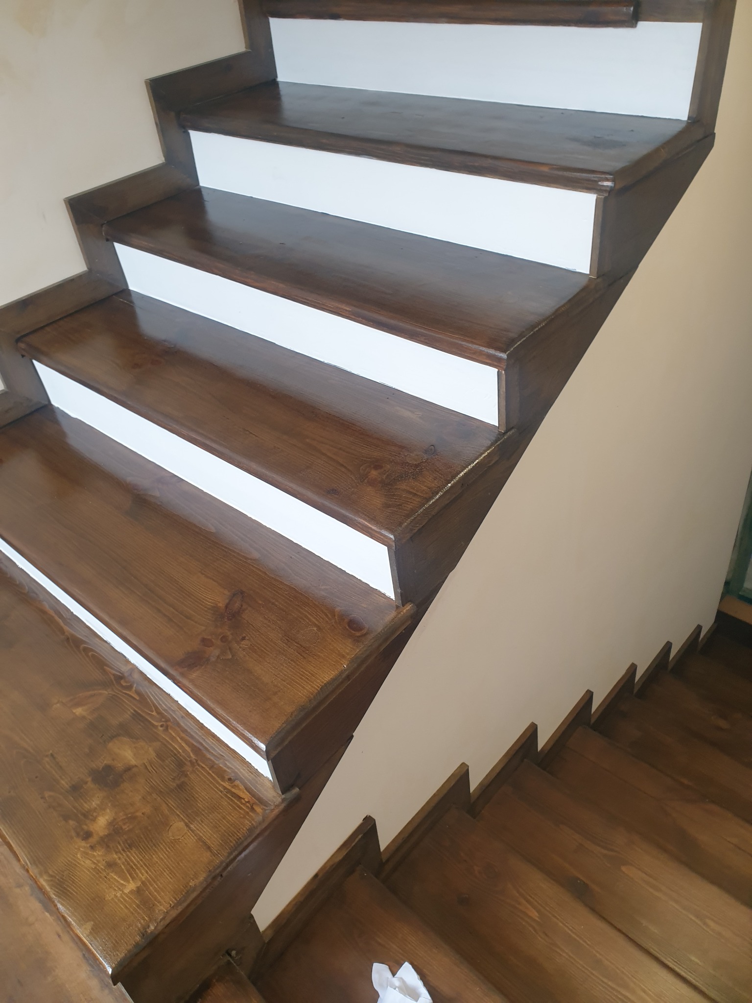 Odnowione drewniane schody z brązowymi stopniami i białymi podstopnicami, widoczne z dołu, z wyraźnym rysunkiem drewna i połyskiem lakieru.