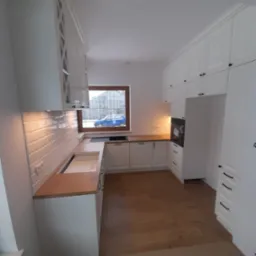 Wnętrze jasnej kuchni z białymi szafkami z frezowanymi frontami i drewnianym blatem, widok na okno z niebieskim samochodem na zewnątrz, drewniana podłoga.