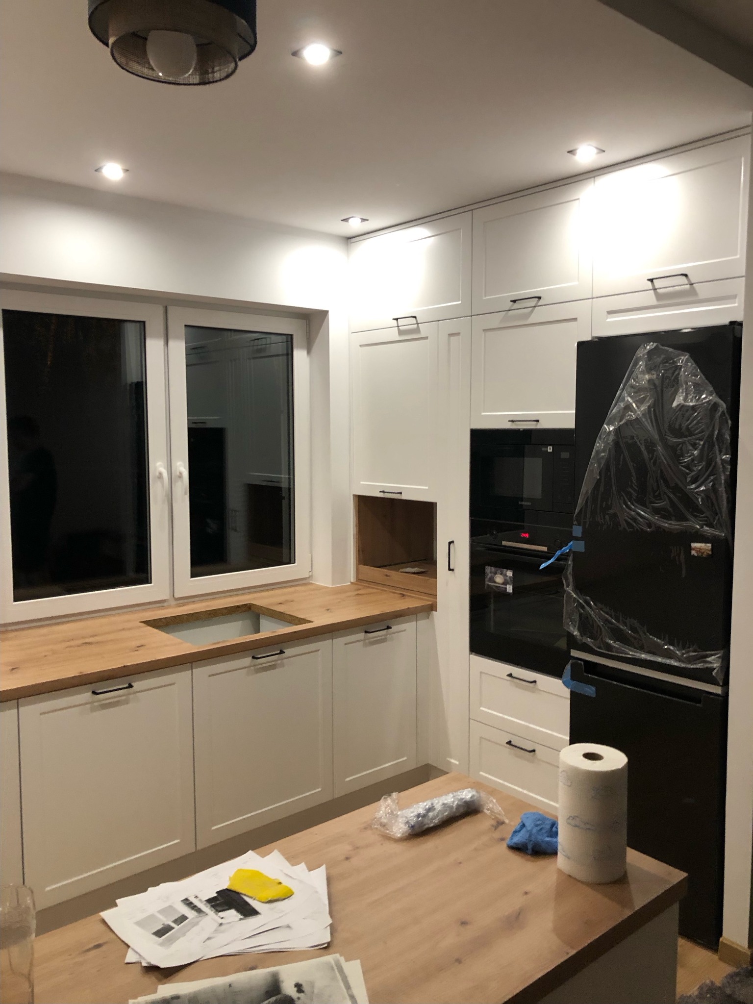 Nowoczesna kuchnia w trakcie montażu: białe szafki z drewnianym blatem, czarny sprzęt AGD jeszcze w folii ochronnej, widoczne plany i akcesoria montażowe na wyspie kuchennej.