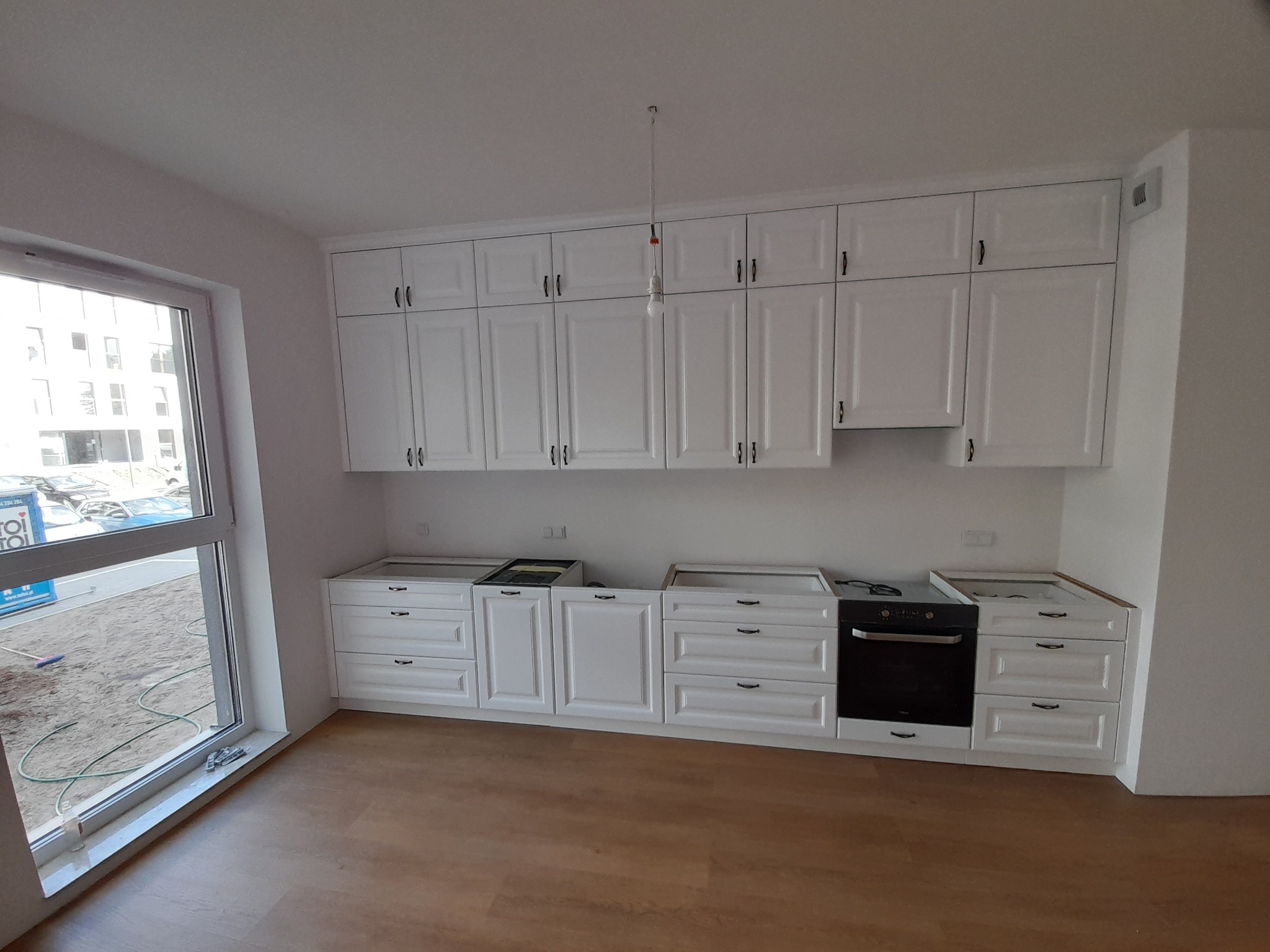 Biała zabudowa kuchenna w trakcie montażu, widoczne niedokończone blaty i wisząca żarówka, parkiet na podłodze.