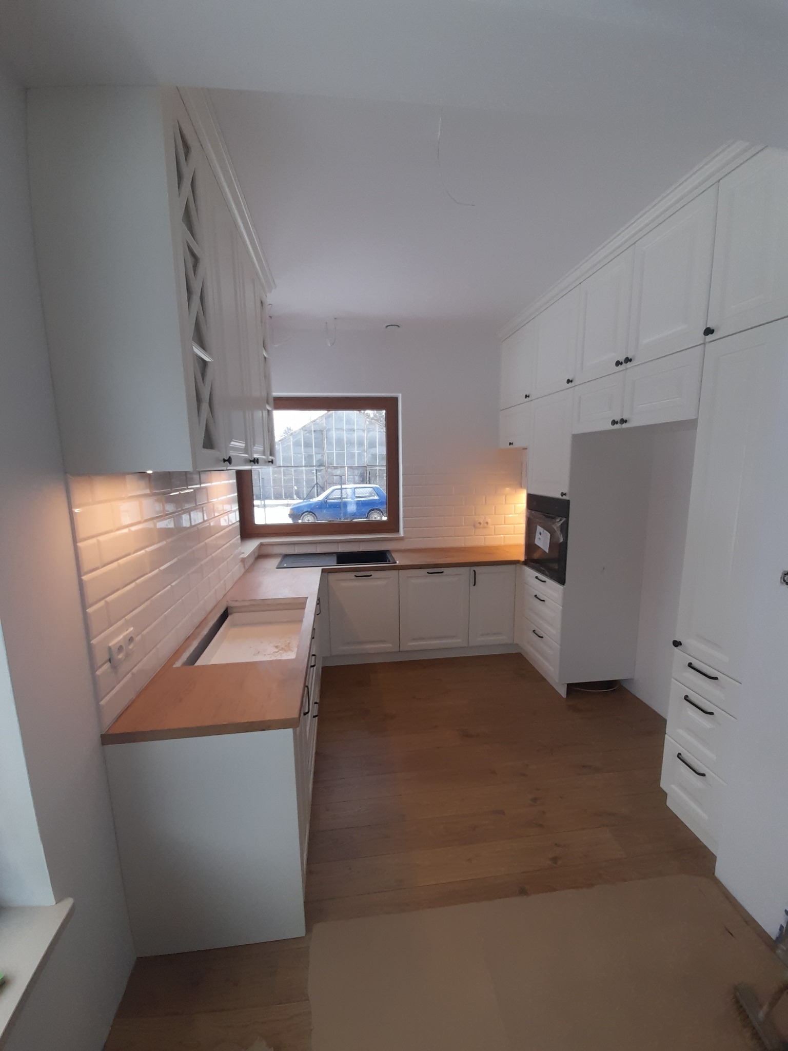 Wnętrze jasnej kuchni z białymi szafkami z frezowanymi frontami i drewnianym blatem, widok na okno z niebieskim samochodem na zewnątrz, drewniana podłoga.