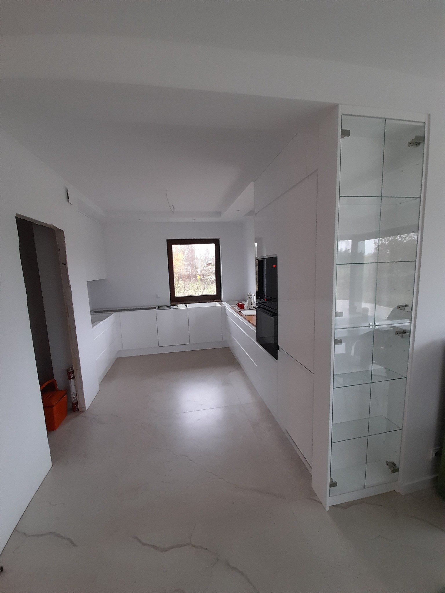 Nowoczesna, minimalistyczna kuchnia z białymi frontami, szklaną witryną i oknem z widokiem na zieleń, jeszcze w trakcie wykończenia, widoczna skrzynka narzędziowa przy ścianie.