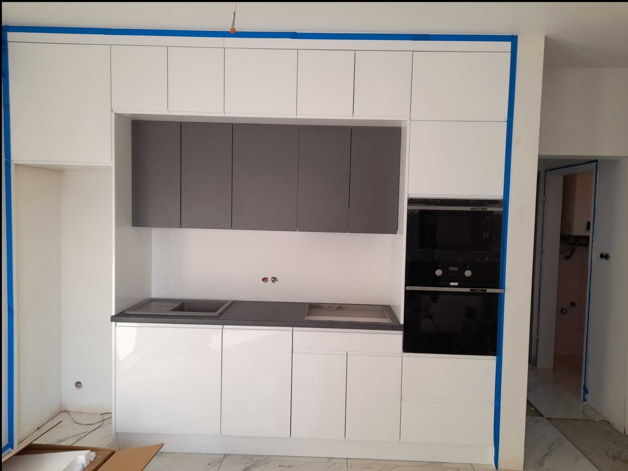 Nowoczesna kuchnia w trakcie montażu: białe szafki górne i dolne, szare szafki wiszące, czarny piekarnik, marmurowe płytki na podłodze, zabezpieczenia z niebieskiej taśmy malarskiej na ścianach.