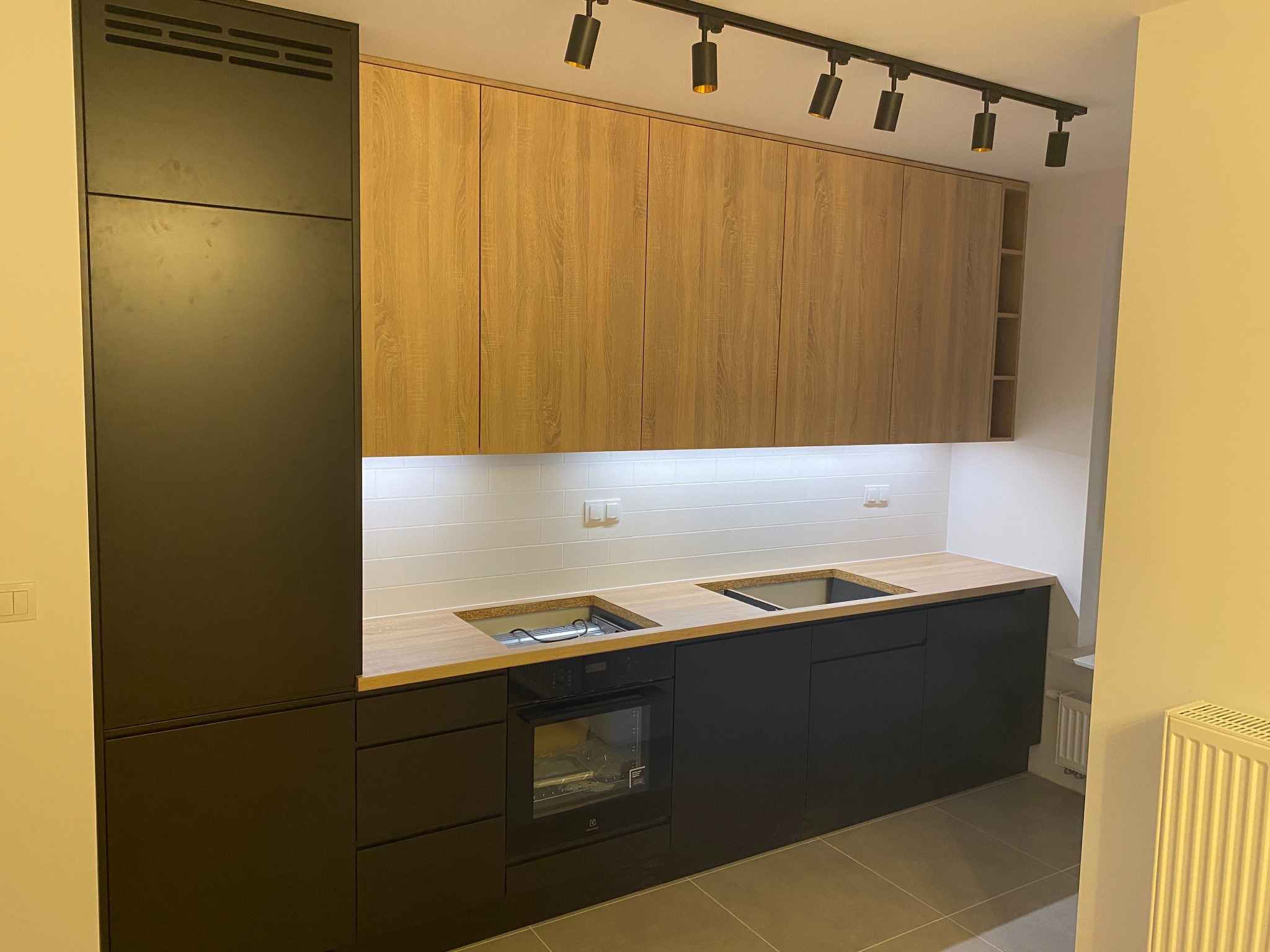 Nowoczesna kuchnia z czarnymi szafkami dolnymi i drewnopodobnymi górnymi, oświetlenie szynowe, białe płytki na ścianie, bez uchwytów, widoczny piekarnik Electrolux.