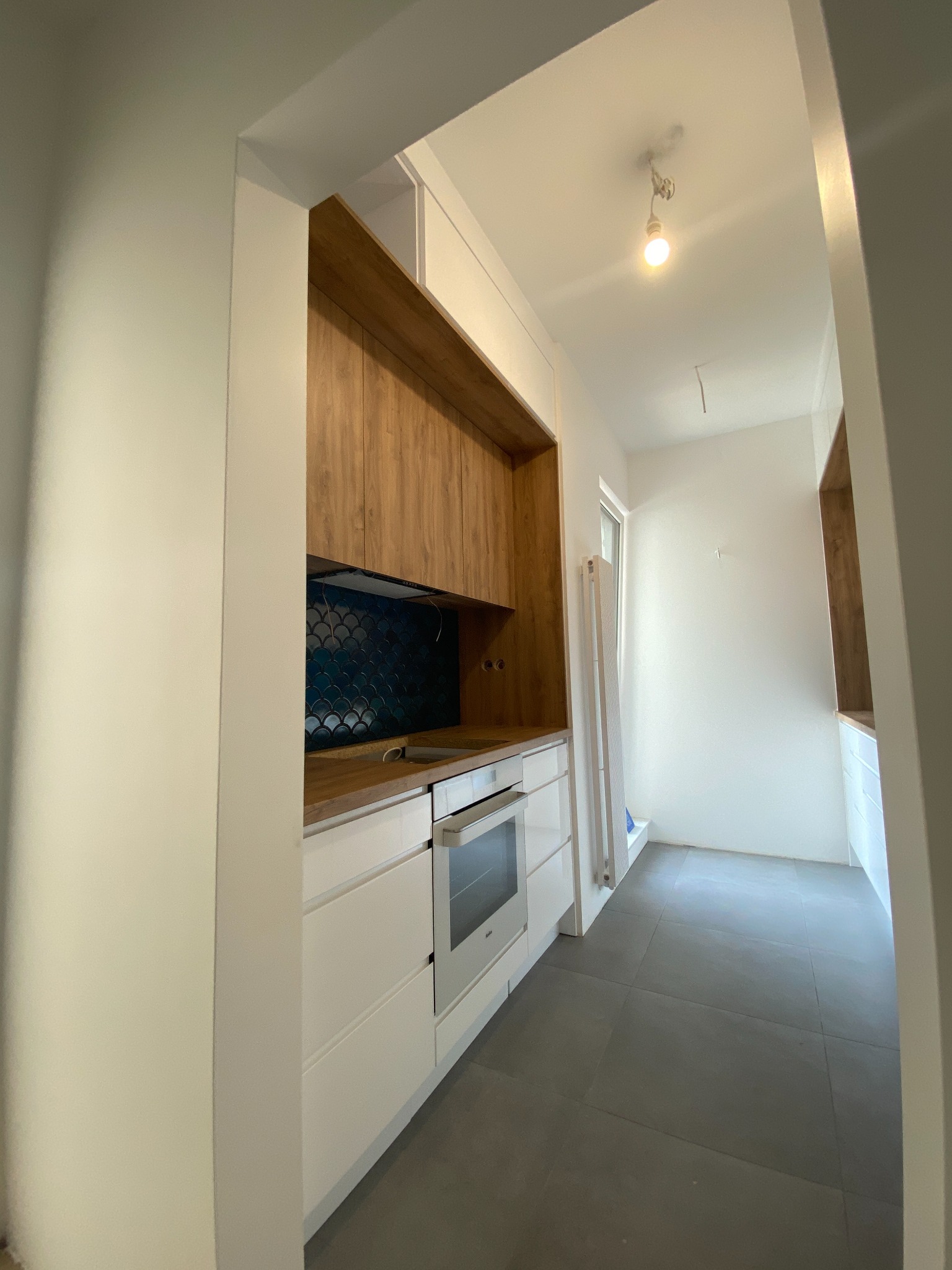 Wnętrze kuchni w trakcie wykańczania: białe szafki dolne, drewniane szafki górne, niebieska mozaika na ścianie, surowa żarówka, szare płytki podłogowe.