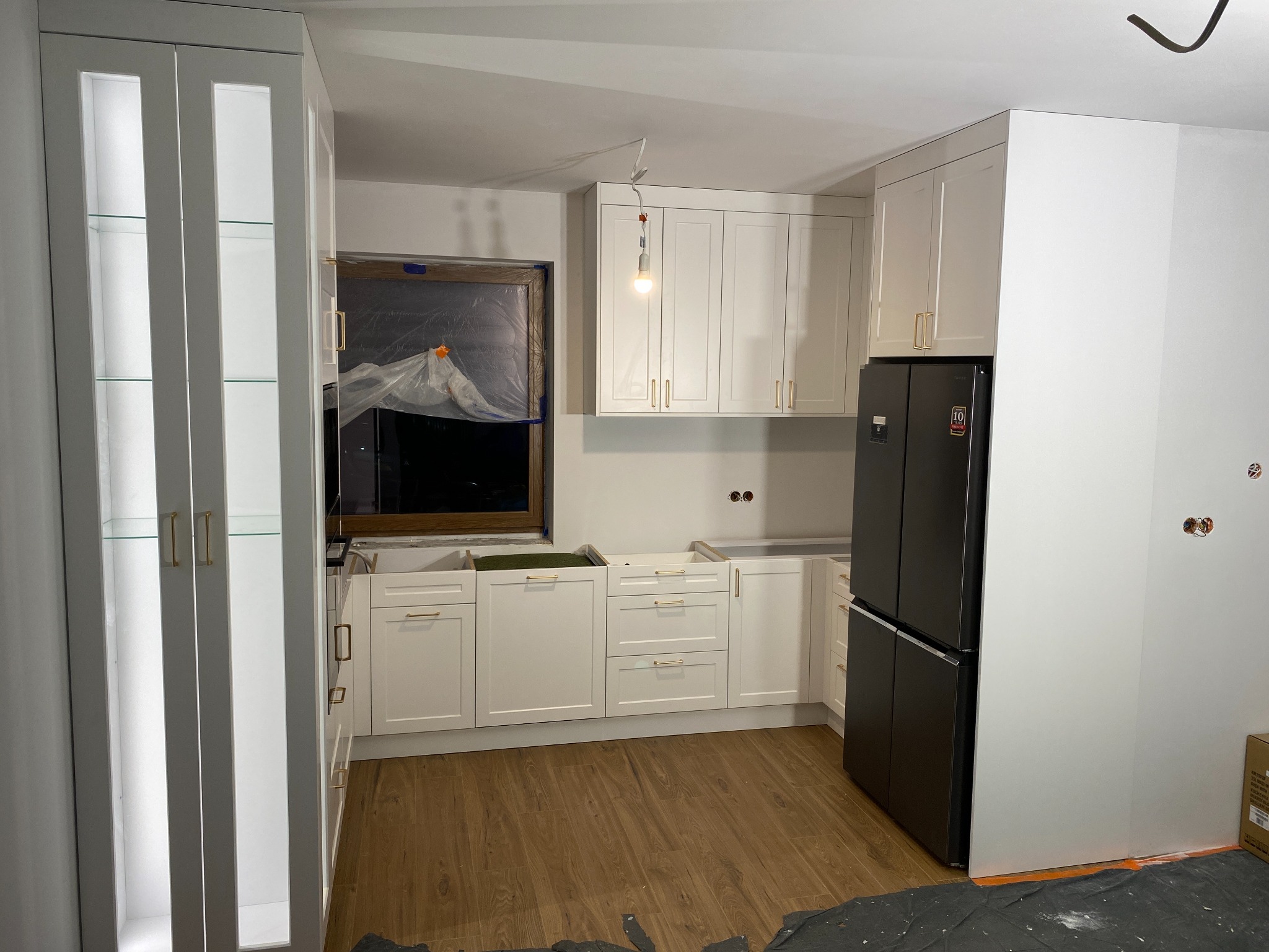 Montaż białej kuchni z szafkami dolnymi i wiszącymi z złotymi uchwytami, widoczna lodówka, okno zabezpieczone folią, surowa żarówka na kablu, podłoga drewnopodobna, etap prac wykończeniowych.
