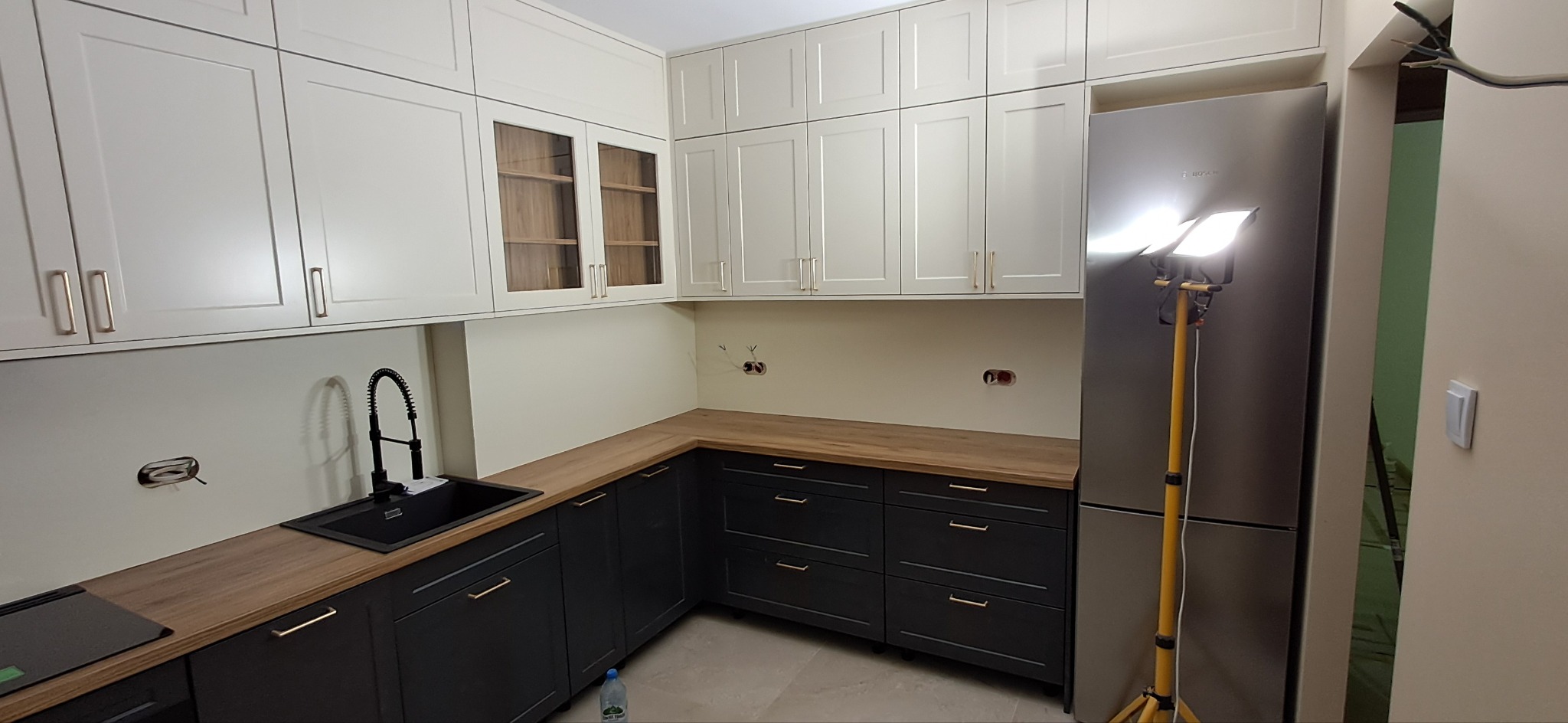 Nowoczesna kuchnia z białymi szafkami wiszącymi i grafitowymi szafkami dolnymi z drewnianym blatem, czarnym zlewem i baterią, oświetlona lampą budowlaną, widoczne gniazdka elektryczne w ścianie.