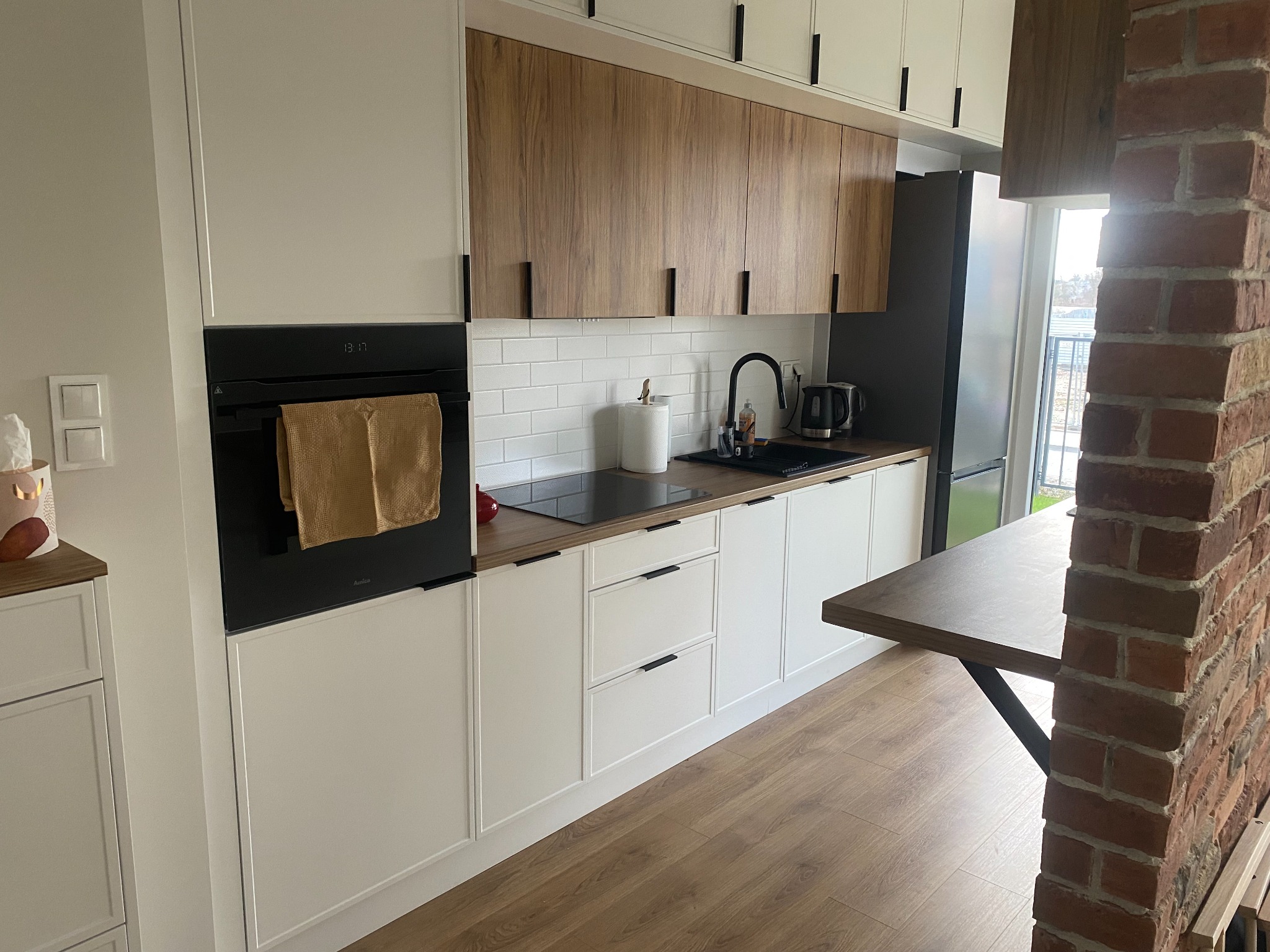 Nowoczesna kuchnia z białymi szafkami dolnymi i drewnianymi górnymi, czarnymi uchwytami, ceglanym filarem i blatem zintegrowanym ze stołem.