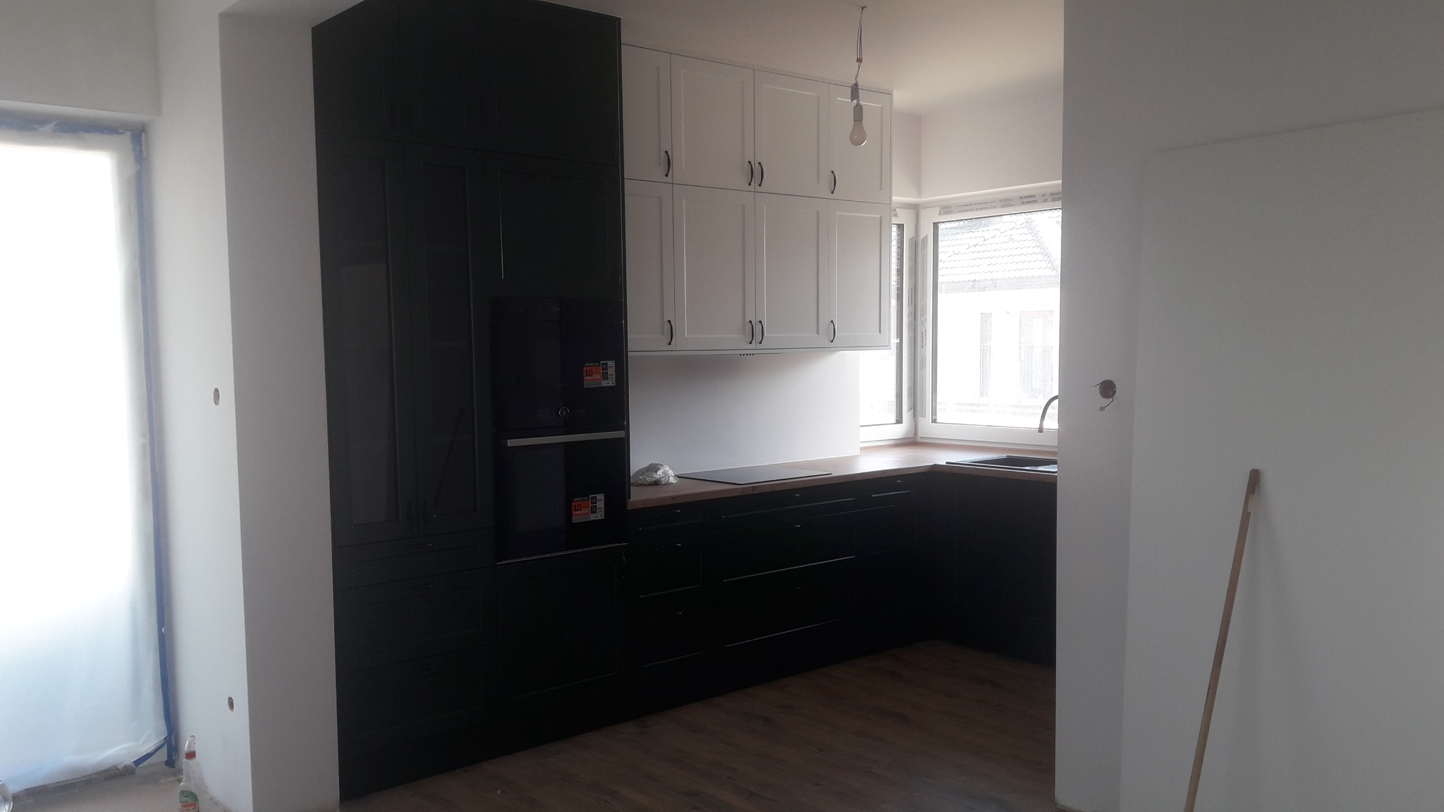 Częściowo wykończona kuchnia z czarnymi dolnymi szafkami i białymi górnymi, z widocznym oknem i niedokończonymi ścianami.