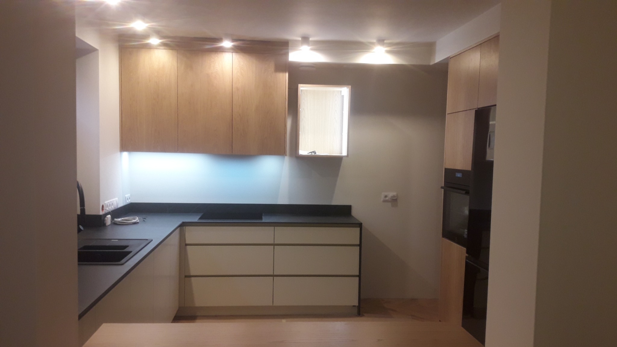 Nowoczesna kuchnia z drewnianymi szafkami górnymi i białymi dolnymi szufladami, czarny blat, oświetlenie LED pod szafkami, zabudowany piekarnik.