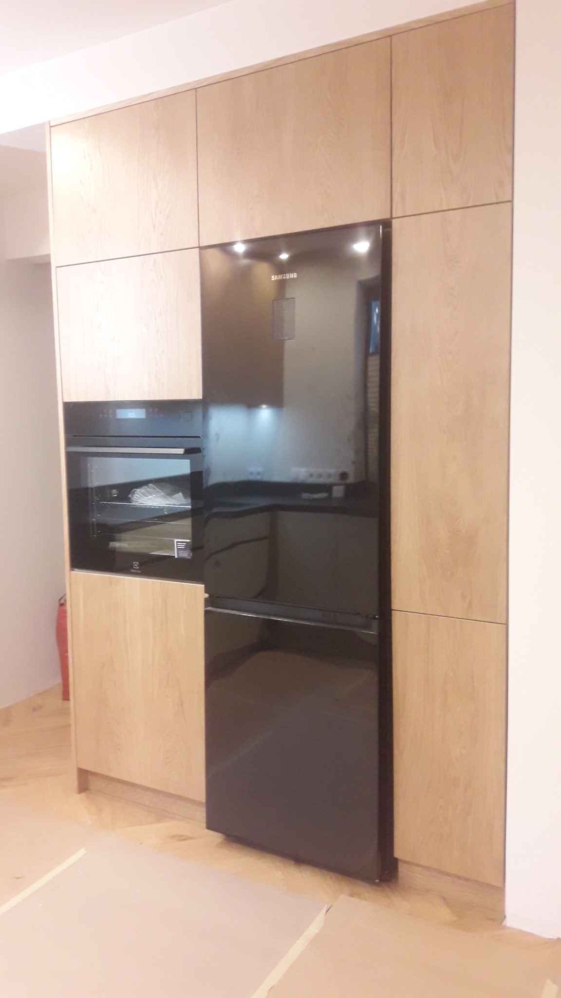 Kuchnia na wymiar: zabudowa z jasnego drewna z wbudowanym piekarnikiem Electrolux i lodówką Samsung z czarnym frontem, widoczne zabezpieczenia podłogi.