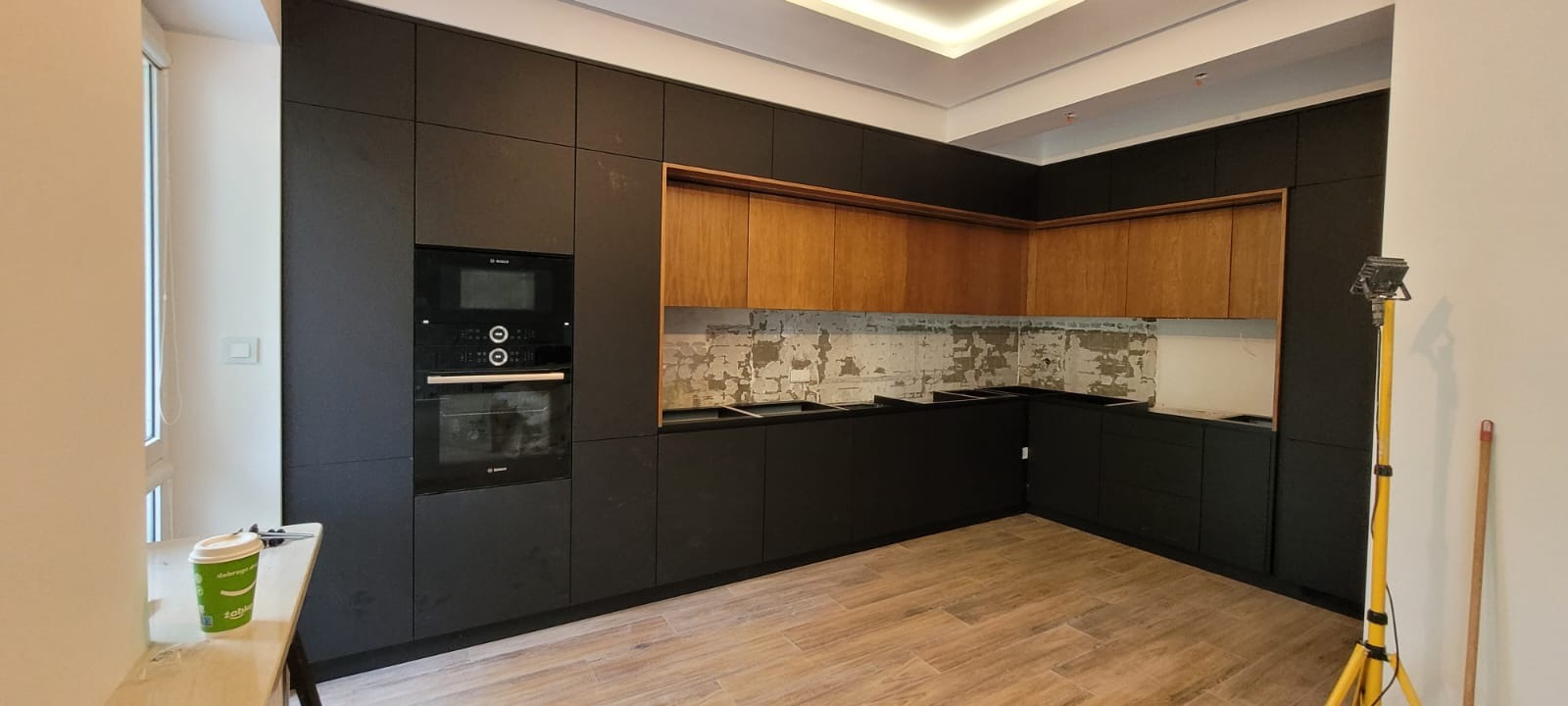 Montaż czarnej zabudowy kuchennej z drewnianymi szafkami wiszącymi i sprzętem AGD w Zielonce, widoczny fragment ściany bez wykończenia i lampa budowlana.