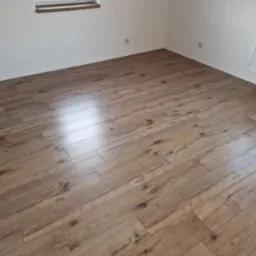 Nowo ułożone, jasnobrązowe panele podłogowe w pustym pokoju z białymi ścianami, widoczne gniazdka elektryczne i grzejnik pod oknem.