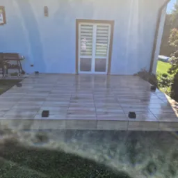 Wykończony taras przed domem, wyłożony płytkami imitującymi drewno. Widoczne czarne podpory pod konstrukcją. W tle ściana budynku i zieleń ogrodu.