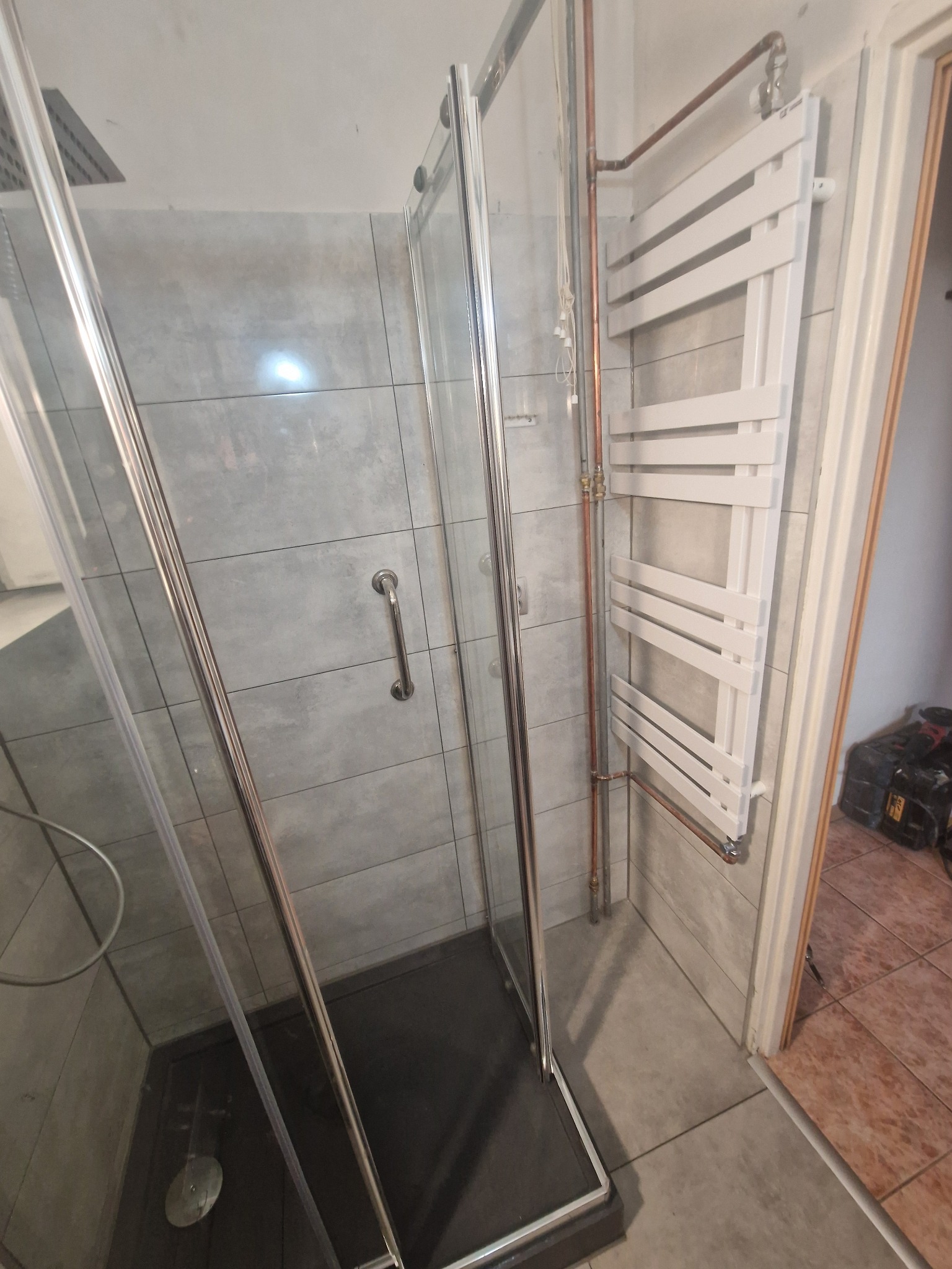 Kabina prysznicowa z szarymi płytkami i grzejnikiem drabinkowym. Miedziane rury doprowadzają wodę do grzejnika. Widoczny fragment podłogi i drzwi.