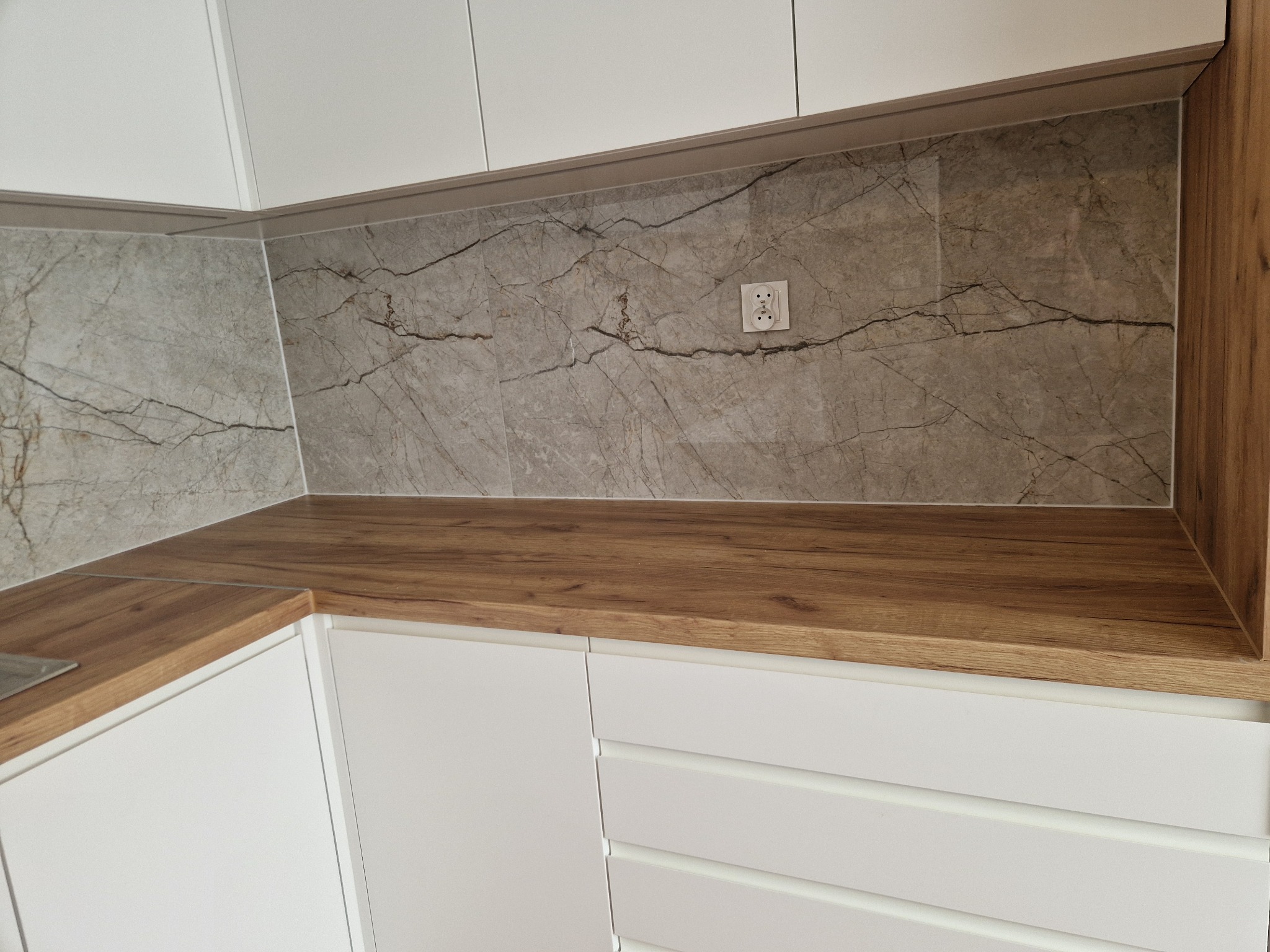 Kuchenny blat z drewnianym dekorem, marmurowe płytki ścienne z gniazdkiem, białe fronty szafek z subtelnymi frezami. Nowoczesne wykończenie kuchni.