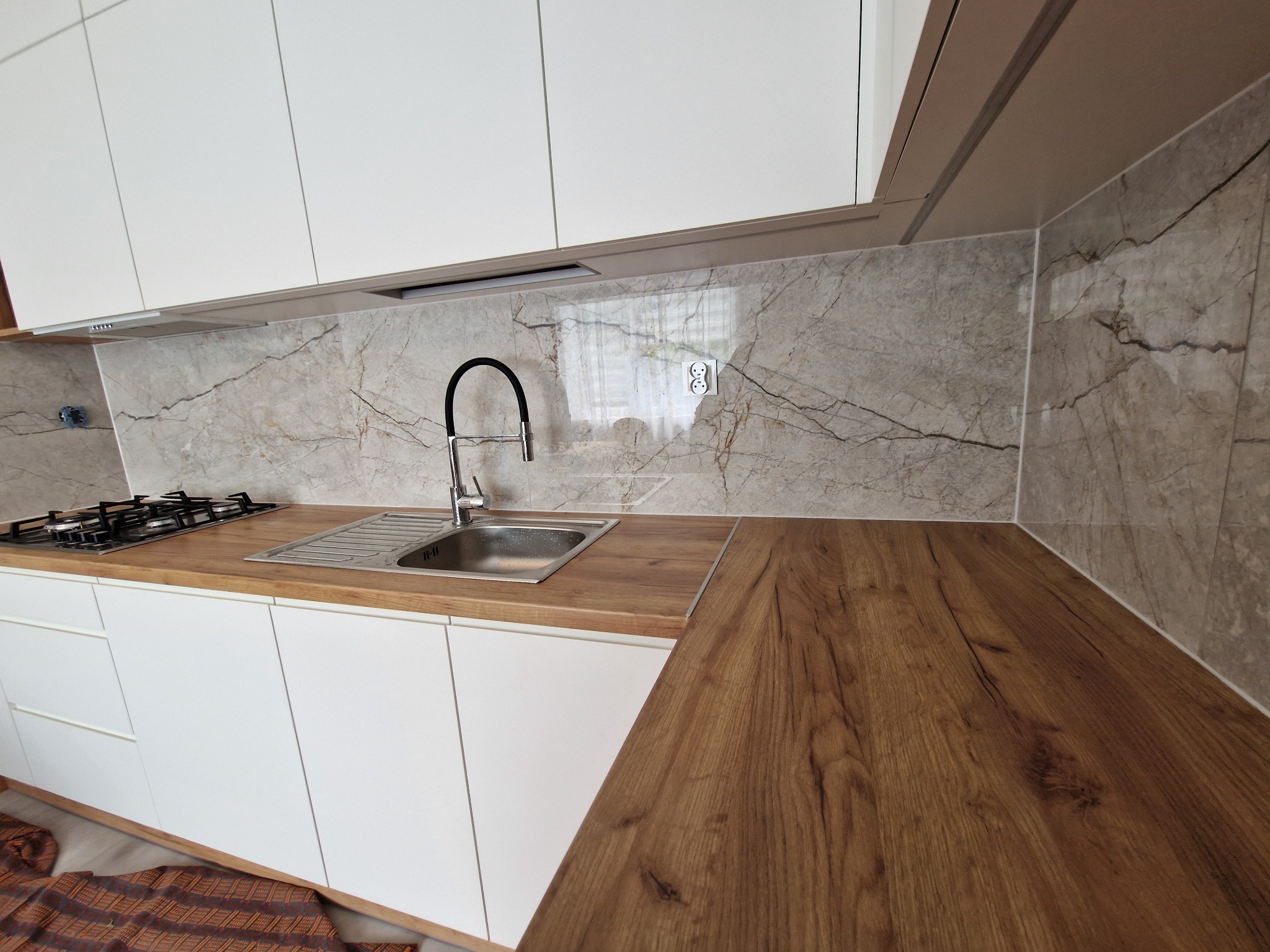 Nowoczesna kuchnia z białymi szafkami, drewnianym blatem i marmurowym panelem ściennym. Widoczna armatura i płyta gazowa.