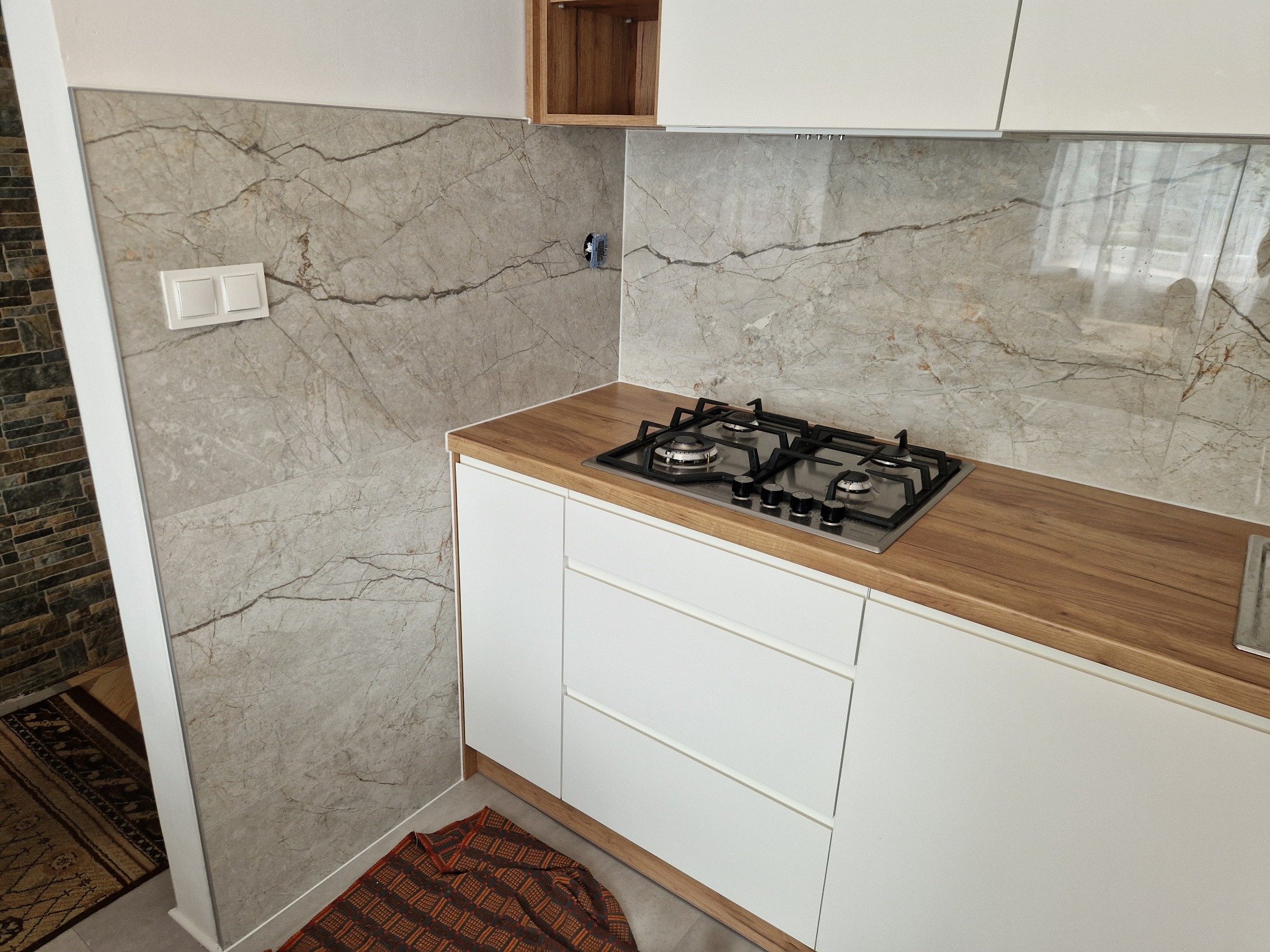 Wykończona kuchnia z płytkami imitującymi marmur na ścianie, białe szafki i drewniany blat. Widoczna płyta gazowa i fragment wejścia do kuchni z dywanem.